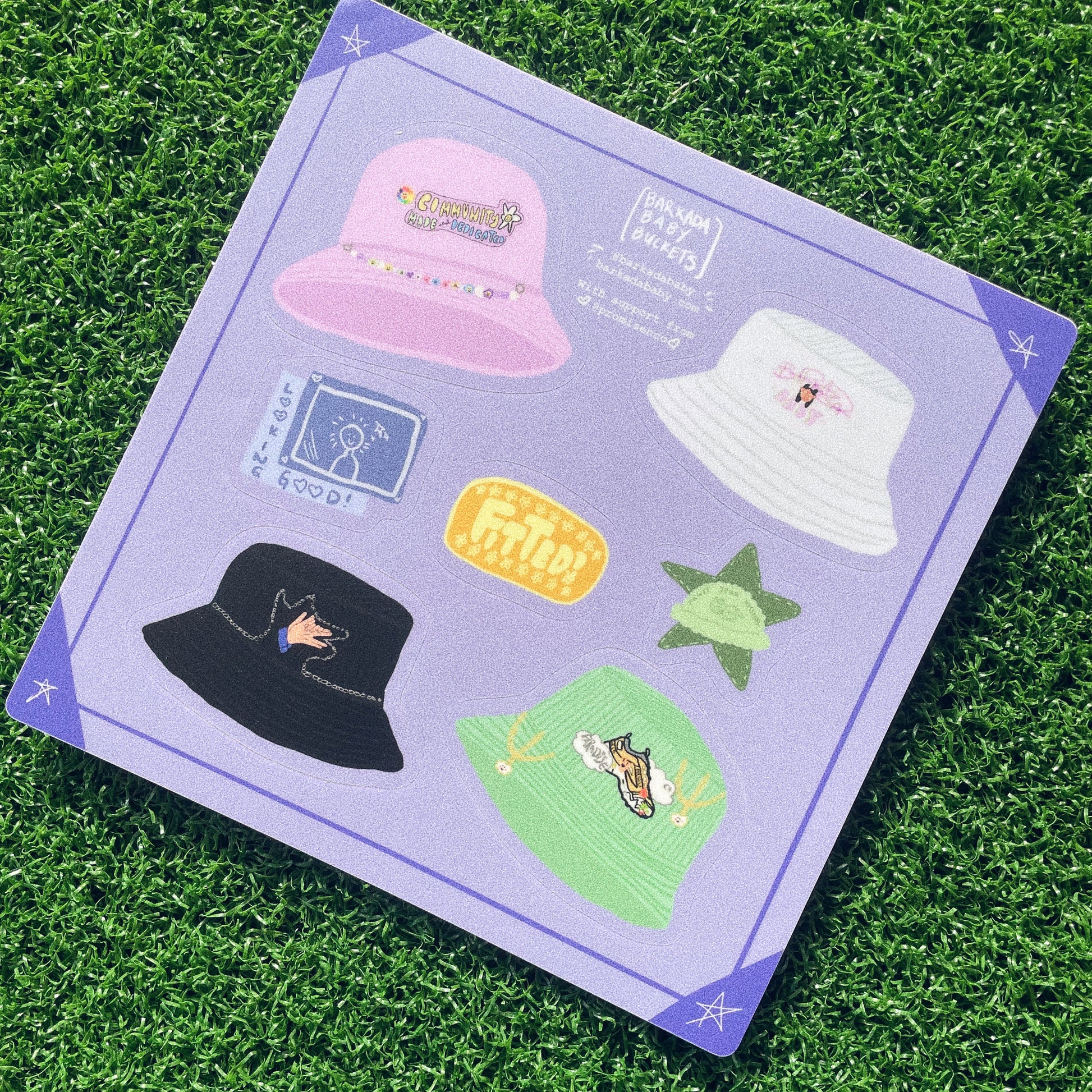 fake grass background with a purple sticker sheet square laying on top. Each sticker is a mini version of a bucket hat made by barkada baby with a "lookin good" sticker, yellow fitted sticker, and a green fuzzy bucket hat sticker with a star in the background