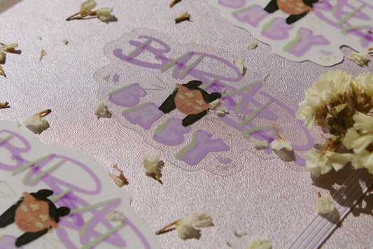purple journal in the background with dried white flowers scattered about. In the middle is a clear sticker and on the top and bottom are the sticker with white backgrounds. The sticker reads "Barkada Baby" in purple lettering with green highlights. In the middle is a person with two buns in their hair, star eyes, and a happy expression. There are lavender and heart details.