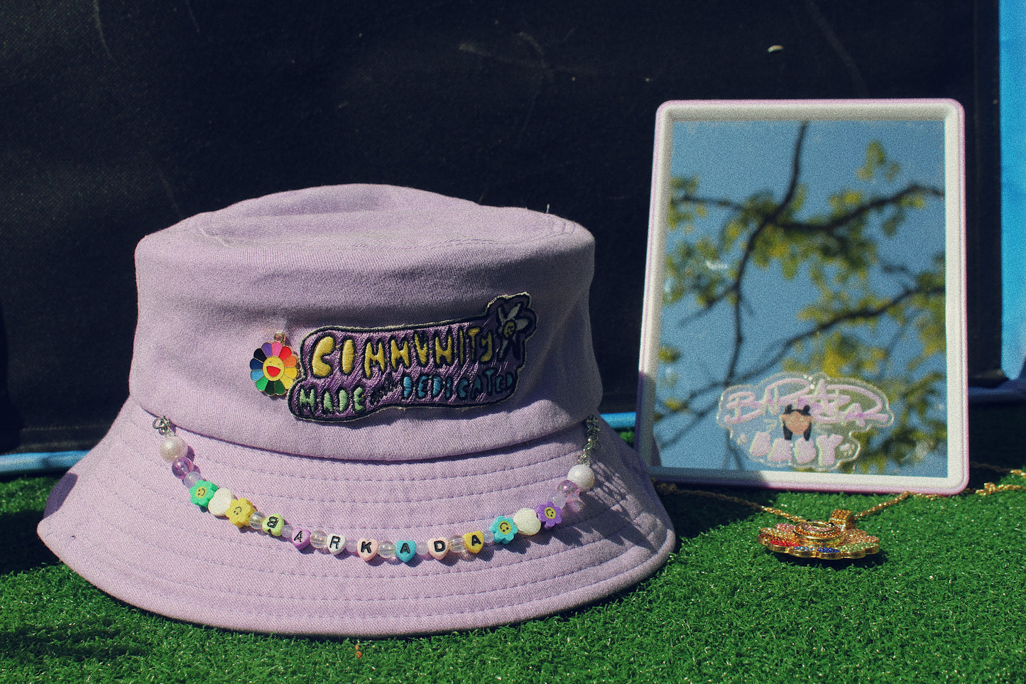 purple community made hat sitting on fake grass next to a mirror and gold necklace