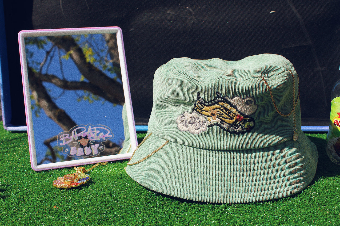 green track 5 paradise hat sitting on fake grass next to a mirror and gold necklace